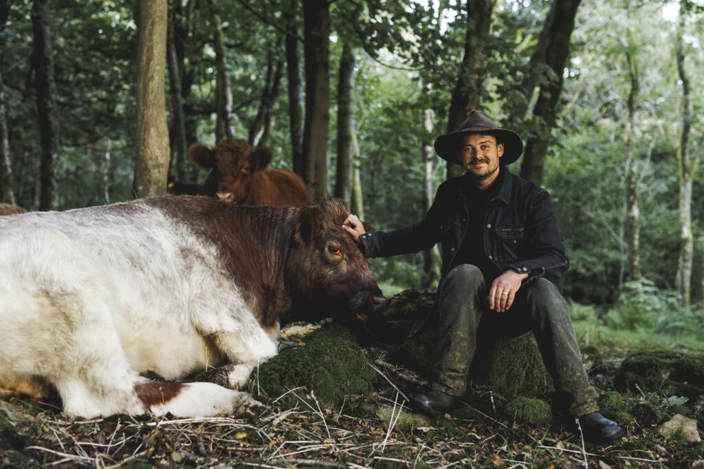 Bobby Keegan sits on a rock in the Lake District countryside with his hand on the head of one of his cows as it lays beside him with another of his cows in the background